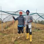 Two smiling farmers prepare the land for a new harvest, with greenhouse tunnels in the background